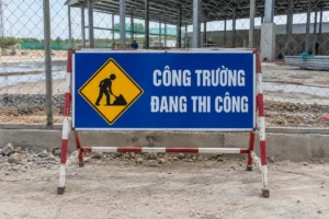 Biển chữ nhật nền xanh Công trường đang thi công, có biểu tượng người đội mũ xúc đất, khung chân chữ A sơn đỏ trắng đặt trước hàng rào lưới
