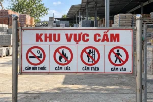 Bảng ngang Khu vực cấm có bốn ký hiệu: cấm hút thuốc, biển cấm lửa, cấm trèo và cấm vào, gắn giữa hai cột inox trước kho vật liệu