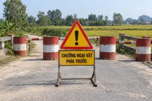 Biển tam giác vàng viền đỏ có dấu chấm than đen đặt trên bảng vàng chữ đen Chướng ngại vật phía trước, giữa đường quê