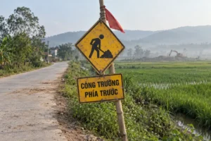 Biển vàng hình thoi có hình người xúc đất, bên dưới là bảng vàng chữ Công trường phía trước, buộc trên cột tre cạnh đường quê