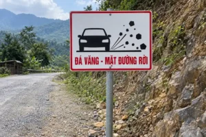 Biển chữ nhật nền trắng viền đỏ có hình ô tô và những viên đá văng sau bánh, dòng chữ đỏ Đá văng - mặt đường rời, gắn trên cột thép cạnh đường đá dăm