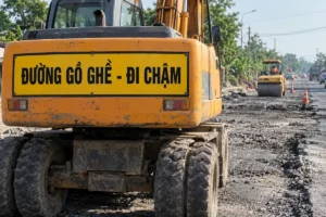 Bảng chữ nhật nền vàng chữ đen Đường gồ ghề - Đi chậm gắn trên thân sau của máy xúc bánh lốp, phía sau là xe lu và mặt đường nhựa đang cào bóc