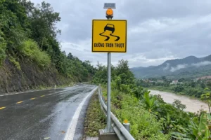 Biển báo đường trơn trượt, biển vuông nền vàng viền đen có hình chiếc xe trượt trên vệt đường uốn lượn và dòng chữ Đường trơn trượt, gắn trên cột thép bên đường núi ẩm ướt, có đèn cảnh báo và tấm pin trên đỉnh cột