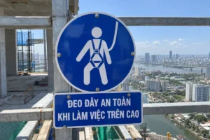Biển báo an toàn lao động bắt buộc đeo dây an toàn: biển tròn nền xanh dương có hình người đeo dây đai toàn thân, bảng chữ ĐEO DÂY AN TOÀN KHI LÀM VIỆC TRÊN CAO, gắn trên thanh thép giàn giáo ở tầng cao