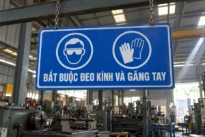 Biển báo an toàn lao động bắt buộc đeo kính và găng tay: biển chữ nhật nền xanh dương có hai vòng tròn hình kính bảo hộ và găng tay, chữ BẮT BUỘC ĐEO KÍNH VÀ GĂNG TAY, treo trong nhà xưởng cơ khí