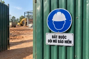 Biển báo an toàn lao động bắt buộc đội mũ bảo hiểm: biển tròn nền xanh dương có hình đầu người đội mũ trắng, bảng chữ BẮT BUỘC ĐỘI MŨ BẢO HIỂM, gắn trên tấm tôn xanh lục cạnh cổng công trường