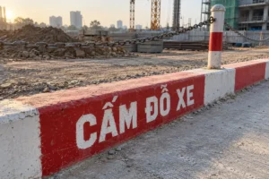 Chữ Cấm đỗ xe sơn trắng trên nền đỏ ở mép bó vỉa sơn đỏ trắng, phía sau có cột và dây xích, công trường đang thi công