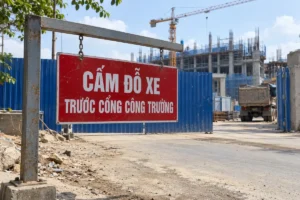 Biển chữ nhật nền đỏ chữ trắng Cấm đỗ xe trước cổng công trường, treo bằng xích trên khung thép cạnh cổng rào xanh
