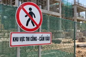 Biển tròn viền đỏ có hình người đi bộ bị gạch chéo, bên dưới là bảng trắng chữ đen Khu vực thi công - Cấm vào, gắn trên hai cột tròn cạnh hàng rào lưới xanh