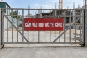 Biển chữ nhật nền đỏ chữ trắng Cấm vào khu vực thi công, treo bằng hai sợi xích trên cổng lưới thép, phía sau là công trình đang xây và cần trục