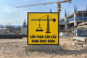 Biển cảnh báo nguy hiểm cần cẩu: biển vuông vàng viền đen có hình cần cẩu tháp treo tải, đặt trên bệ bê tông cạnh công trường