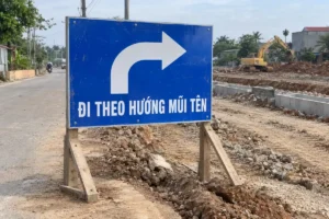 Bảng chữ nhật nền xanh mũi tên cong trắng và chữ Đi theo hướng mũi tên, đặt cạnh đường đang thi công