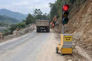 Đèn tín hiệu tạm thời đặt trên xe đẩy vàng có bảng chữ Đèn tín hiệu tạm thời, cạnh đoạn đường đang xử lý đất đá, có xe tải và máy xúc