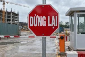 Biển bát giác nền đỏ viền trắng chữ Dừng lại gắn trên cột thép, phía sau là chòi bảo vệ, cổng công trường và cần trục