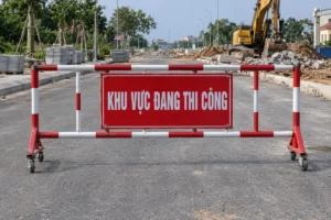 Biển nền đỏ chữ trắng Khu vực đang thi công gắn giữa khung rào sắt sơn đỏ trắng có bánh xe, chắn ngang đường đang làm