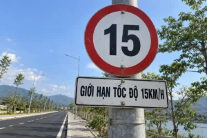 Biển báo giới hạn tốc độ hình tròn viền đỏ nền trắng ghi số 15, bên dưới có bảng phụ chữ Giới hạn tốc độ 15km/h, gắn trên cột thép cạnh con đường ven sông