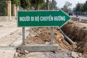 Biển hình mũi tên nền xanh lục chữ trắng Người đi bộ chuyển hướng, đặt cạnh rãnh đào trên vỉa hè đang thi công
