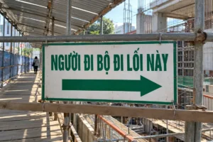 Bảng nền trắng viền xanh chữ Người đi bộ đi lối này, mũi tên sang phải, gắn trên giàn giáo cạnh lối đi có mái che