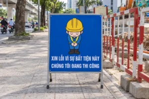 Biển xin lỗi vì sự bất tiện này và Chúng tôi đang thi công, nền xanh chữ trắng, có hình người đội mũ bảo hộ cúi chào, đặt trên vỉa hè cạnh hàng rào đỏ trắng